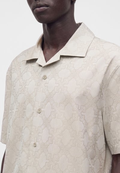 Homme portant une chemise beige à manches courtes avec un motif géométrique subtil et un col camp.