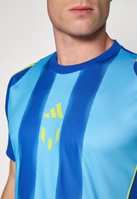 Blå atletisk trøje med skiftende lyse og mørkeblå striber. Designet inkluderer gule accenter og et logo på brystet.