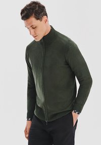 Ciemnozielony sweter na zamek z wysokim kołnierzem. Dzianinowy materiał, ściągacze na rękawach i unikalne wzorzyste akcenty na wewnętrznych mankietach. Dopasowany krój.