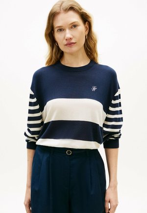 Kvinde med bølget blondt hår iført marineblå og hvide striber på en langærmet sweater og marineblå bukser med høj talje og guldspænde.