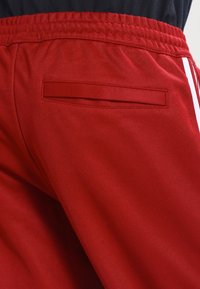 Červené sportovní kalhoty s elastickým pasem, plochou zadní kapsou a bílými pruhy na bocích. Látka vypadá hladká a odolná.