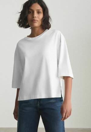 Femme aux cheveux foncés mi-longs, portant une chemise blanche ample à manches trois-quarts et un jean bleu devant un fond uni.