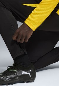 Sorte atletiske bukser med en lynlås lomme, parret med sorte Puma støvler med en tekstureret overflade og hvide logo accenter. Gul ærme synlig.