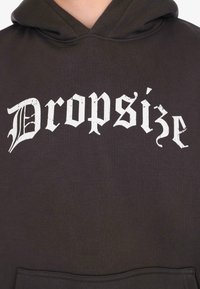 Nahaufnahme eines dunklen Kapuzenpullovers mit weißem, gotischem Schriftzug "Dropsize" auf der Brust sowie einem Teil der Kängurutasche sichtbar.