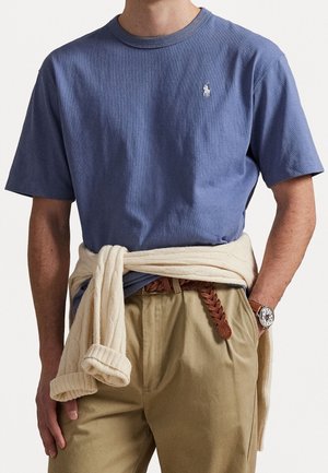 Mand iført en blå kortærmet skjorte, beige bukser med flettet læderbælte, ur og cremefarvet sweater bundet omkring livet.