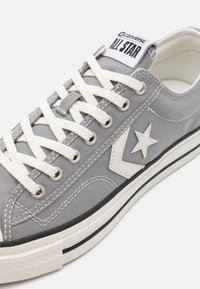 Sneaker in tela grigia con lacci bianchi, suola in gomma bianca e decorazione a forma di stella sul lato. Include un'etichetta con logo bianca e nera.