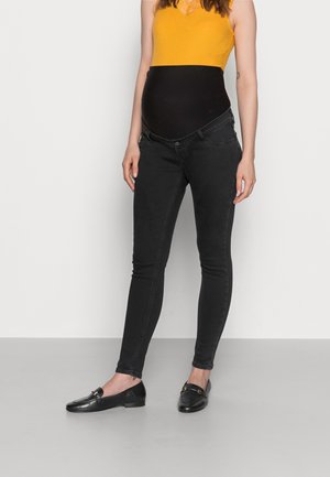 Zwangere persoon in een gele mouwloze top en zwarte zwangerschapsjeans met elastische tailleband en zwarte instappers, staande op een grijze vloer.