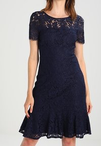 Donna che indossa un vestito in pizzo blu navy aderente con maniche corte e un orlo a volant, in piedi davanti a uno sfondo neutro.