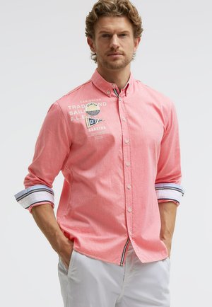 Mann mit lockigem Haar, der ein korallenfarbenes Hemd mit nautischen Aufnähern und aufgekrempelten Ärmeln trägt und mit den Händen in den Taschen einer weißen Hose steht.