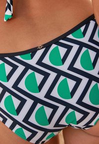 Bikini kalhotky s geometrickým vzorem v zelené a bílé, lemované námořnickým modrým. Vyznačují se jemným zlatým kovovým detailem poblíž pásku.