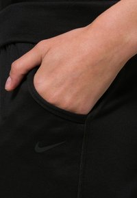 Pantalones deportivos negros con un bolsillo lateral; incluyen un logo tonal de Nike. La tela parece suave con un ligero estiramiento y una cintura ajustada.