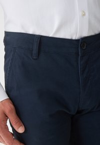 Námornícky modré bavlnené chinos nohavice s hladkou štruktúrou, vybavené gombíkovým zapínaním a pútkami na opasok. Na vrchu je čiastočne vidieť biele tričko.