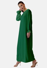 Długa zielona sukienka z gładkiego materiału, z bufkami na długich rękawach i luźną sylwetką. Noszona z przezroczystymi sandałami na obcasie.