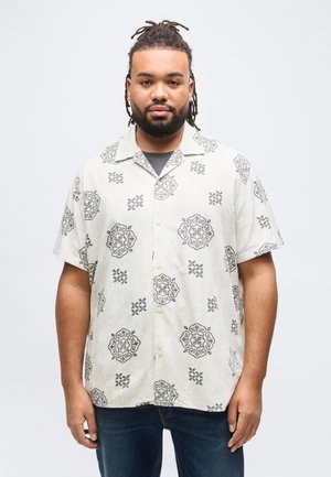 Man with beard and dreadlocks wearing a short-sleeve white shirt with black floral patterns, standing against a plain light background.