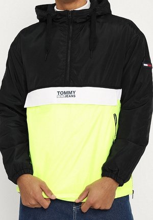 Mand iført en sort og neongul Tommy Jeans hættetrøje med halvlukning og logo på tværs af brystet.