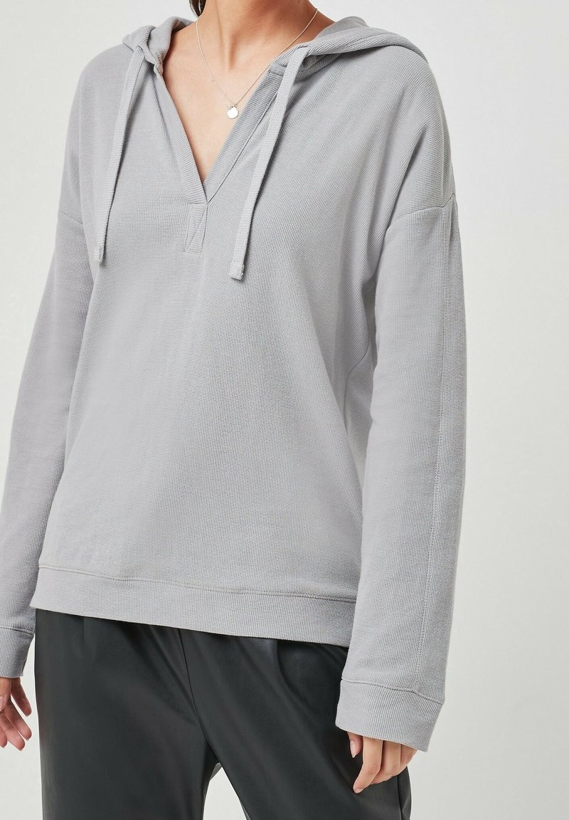 Šedá texturovaná mikina s výstřihem do V, šňůrkami a širokými rukávy. Kombinováno s černými kalhotami, které mají uvolněný střih a ležérní design.