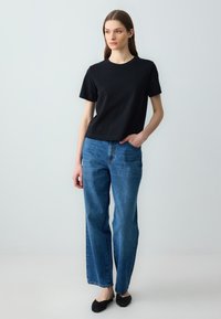 Kobieta stoi z jedną ręką w kieszeni niebieskich dżinsów, ubrana w czarną koszulę z krótkim rękawem i czarne buty na slip-on, na neutralnym tle.