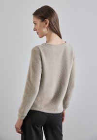Maglione beige peloso con maniche lunghe e scollatura arrotondata, abbinato a pantaloni in tessuto scuro. Texture liscia e design semplice.