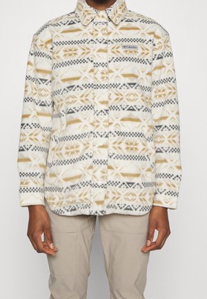 Persoon draagt een beige, zwarte en witte fleecejas met patroon, een kraag en Columbia-logo op de borst, gecombineerd met lichtbeige broek.