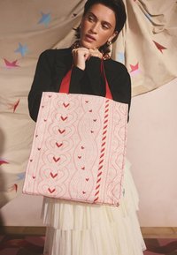 Roosa kangast ostukott punaste detailidega, millel on kootud muster ja südamekujud. Kaks punast sangad, tikandiga tekstuur ja triibuline detail.