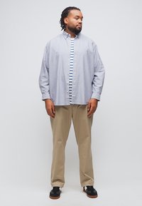 Ενήλικας άνδρας με πλεξούδες που φορά ανοιχτό γαλάζιο πουκάμισο πάνω από ριγέ μπλούζα, μπεζ παντελόνια και μαύρα αθλητικά παπούτσια, στέκεται μπροστά σε απλό φόντο.