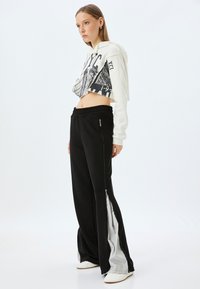 Beschnittene weiße Kapuzenjacke mit grafischem Druck und schwarzen hochgeschnittenen Hosen mit seitlichen Reißverschlüssen und grauen Akzenten. Glatte Stofftextur.