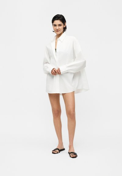 Γυναίκα με κοντά σκούρα μαλλιά, φοράει υπερμεγέθη λευκό πουκάμισο με κουμπιά και μαύρα σανδάλια slide, στέκεται μπροστά σε ένα απλό λευκό φόντο.