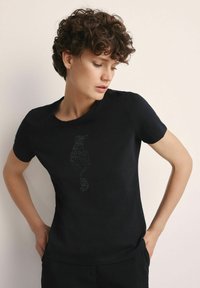 Kobieta z krótkimi kręconymi włosami, ubrana w czarną koszulkę z wzorem ptaka z kamieniami ozdobnymi, patrząca w dół z rękami na biodrach na tle jasnej ściany.