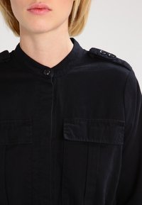 Kobieta ubrana w czarną koszulę z guzikami, z epoletem i dwiema kieszeniami na piersi, ukazana od brody do połowy torsu.