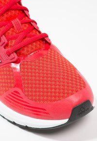 Zapatilla deportiva roja con un empeine de malla texturizada, superposiciones sintéticas lisas y una suela de goma. Presenta cordones planos y un detalle de logo.