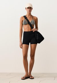 Young woman wearing a patterned bikini top, black high-waisted shorts, beige cap, black crossbody bag, and black flip-flops standing on tiled floor.