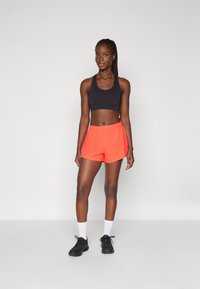 Soutien-gorge de sport noir, short de sport orange avec taille élastique, associé à des chaussettes blanches et des sneakers noirs. Cadre intérieur avec fond neutre.