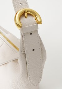 Beige lærveske med stropp og gullmetallakkent. Teksturen er ruglete, med en sømdetalj og en gullglidelås.