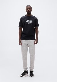 Άνδρας που στέκεται φορώντας μαύρο T-shirt με λογότυπο, ανοιχτό γκρι φόρμα και μαύρα αθλητικά παπούτσια, μπροστά σε απλό λευκό φόντο.