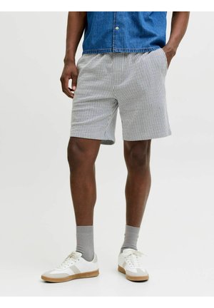 Férfi szürke-fehér csíkos rövidnadrágban, szürke zokniban, fehér tornacipőben és kék farmer ingben, kezét a zsebében tartva.