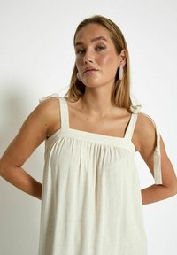 Frau mit blonden Haaren trägt ein ärmelloses cremefarbenes Kleid mit Schulterbändern und großen ovalen Ohrringen und steht vor einem schlichten weißen Hintergrund.