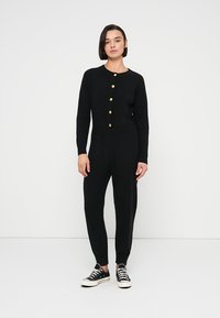 Μαύρη πλεκτή ολόσωμη φόρμα με ριμπ υφή, στρογγυλή λαιμόκοψη και χρυσά κουμπιά ως διακοσμητικά στοιχεία. Διαθέτει εφαρμοστά μανσέτα και κωνικό σχεδιασμό στα πόδια.