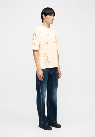 Νεαρός άνδρας που φοράει κρεμ μπλουζάκι με πολύχρωμα κεντημένα σχέδια και σκουρό μπλε τζιν παντελόνι, στέκεται μπροστά σε απλό λευκό φόντο.