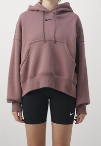 Ylisuuri huppari vaaleanpunaisessa sävyssä, jossa on musta logo, puskuritasku ja resorireunus, yhdistetty mustiin tiukkoihin shortseihin, joissa on logo.