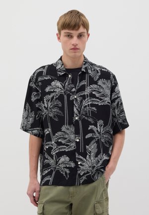 Chemise noire à manches courtes avec un motif blanc de feuilles de palmier, en tissu léger, design col et devant à boutons, associée à un pantalon cargo vert.