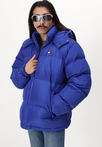 Chaqueta de plumas azul con un diseño acolchado, cierre con cremallera y capucha ajustable. Presenta un pequeño logo en el pecho y puños elásticos.