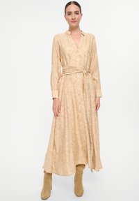 Robe beige à manches longues avec un décolleté en V, motif texturé ondulé, devant à boutons, associée à des bottes beiges montantes. Inclut une ceinture nouée à la taille.