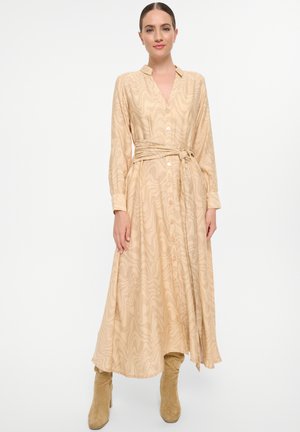 Beige Kleid mit langen Ärmeln und V-Ausschnitt, texturiertem, welligem Muster, Knopfverschluss vorne und kombiniert mit kniehohen braunen Stiefeln. Enthält einen gebundenen Taillengürtel.