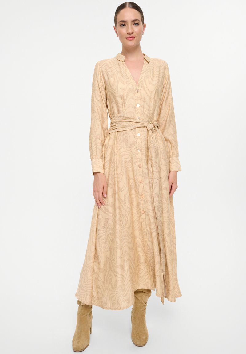 Robe beige à manches longues avec un décolleté en V, motif texturé ondulé, devant à boutons, associée à des bottes beiges montantes. Inclut une ceinture nouée à la taille.