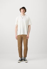 Cream short-sleeve t-shirt, brown slim-fit pants, black high-top sneakers with white soles, and silver accessories.