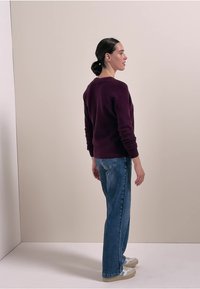 Stickad mörklila tröja, avslappnad passform, rund halsringning; kombinerad med ljusblå rakbukse och beiga sneakers. Modellen står på sidan.