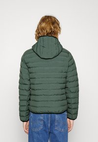 Zelená zateplená bunda s kapucí, která má prošívaný design a vodorovné švy. Tkanina vypadá měkce a má matný povrch.