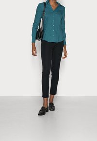 Camicia azzurro scuro con colletto a punta, abbinata a pantaloni neri a tre quarti e mocassini neri. L'outfit include una piccola borsa a tracolla nera.