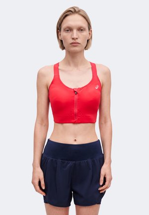 Donna con capelli biondi corti che indossa un reggiseno sportivo rosso con cerniera frontale e pantaloncini sportivi blu scuro, in piedi davanti a uno sfondo semplice.