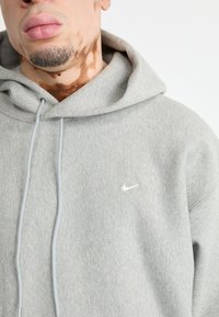 Pelēks Nike kapuce ar mīkstu tekstūru, kas ir aprīkota ar priekšējo kangaroo kabatu, regulējamu auklu un mazu balto logotipu uz kreisā krūtīm.